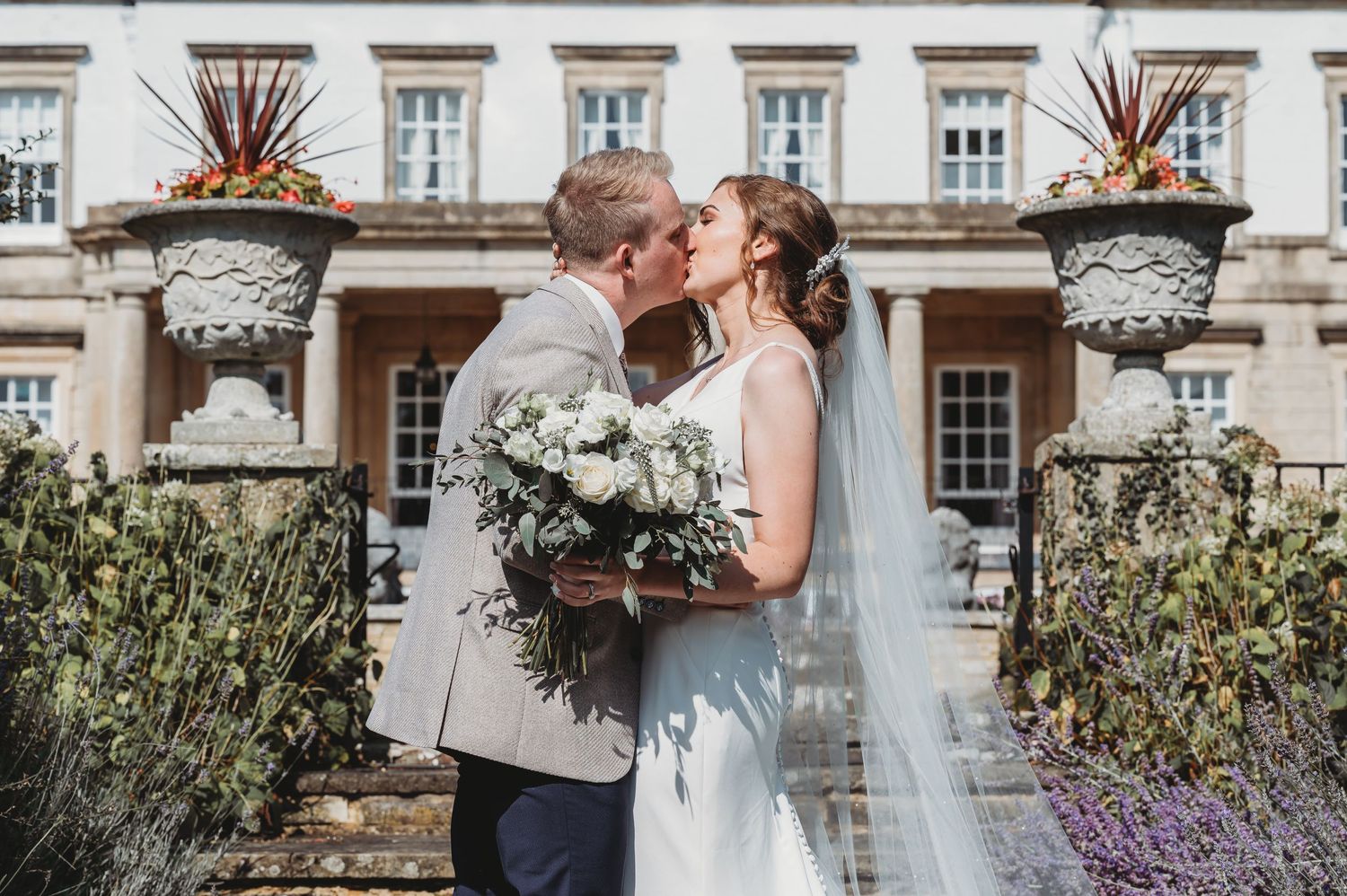 A romantic wedding couple shares a kiss in front of an elegant manor house with ornate potted plants and purple flowers.