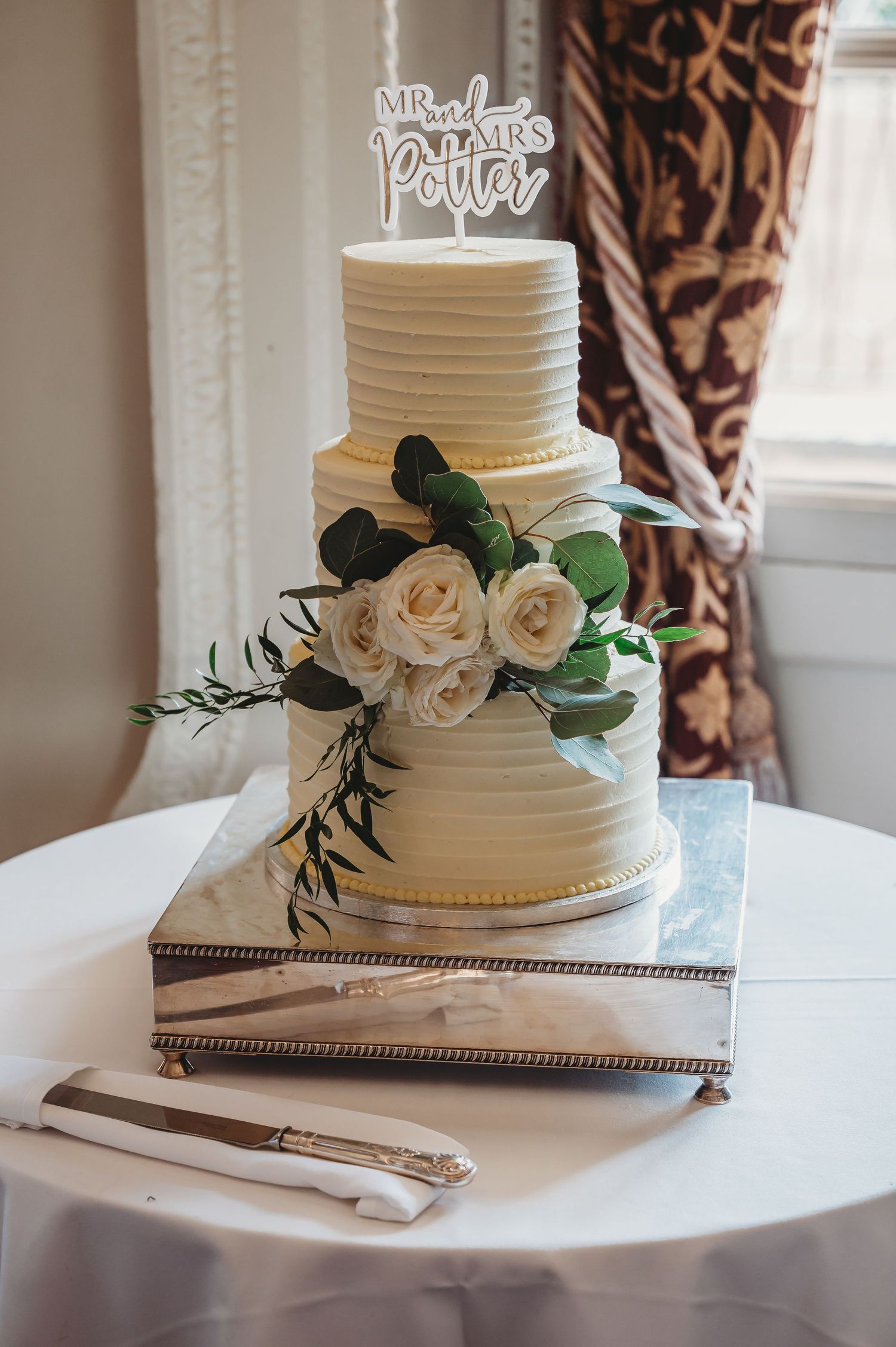 Elegant white two-tier wedding cake with horizontal line texture and white roses on silver platter with cake cutting tools nearby.