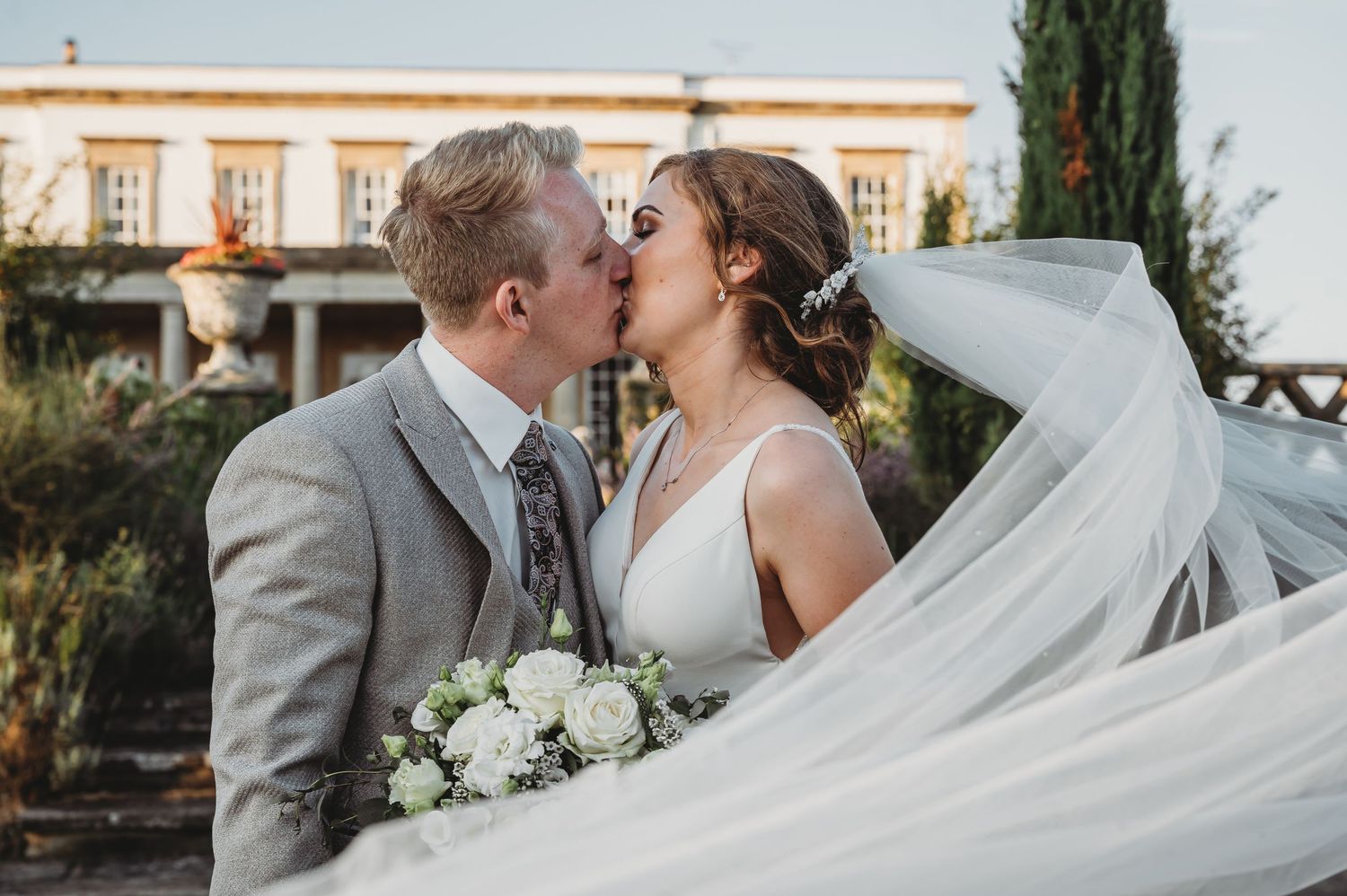 A romantic wedding moment captured at sunset with flowing white veil and bridal bouquet in an outdoor garden setting.