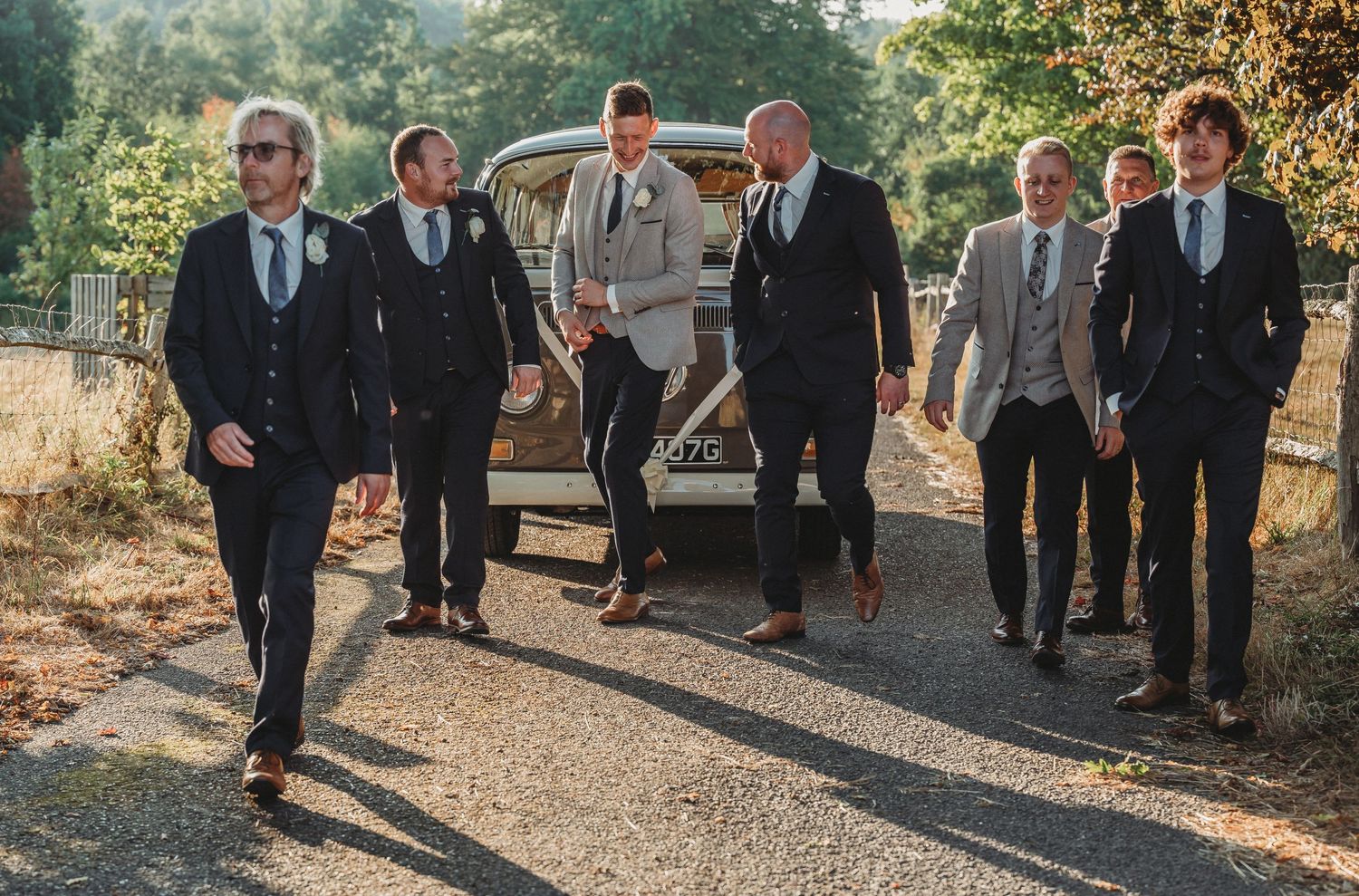 Groomsmen in formal suits walking together on a sunlit path during a wedding celebration.