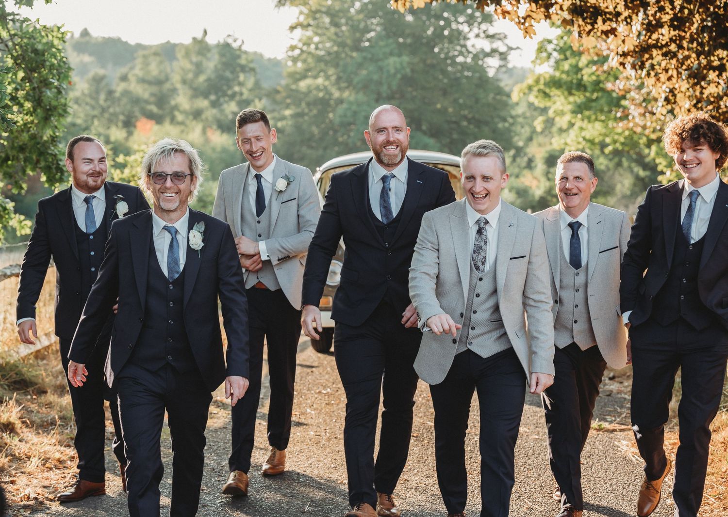 A group of groomsmen in coordinated grey and black suits walking together outdoors at sunset.