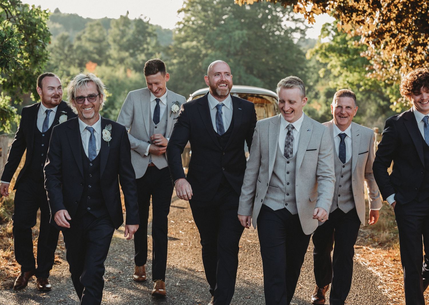 A group of groomsmen in formal suits walking together on a sunlit path during a wedding celebration.