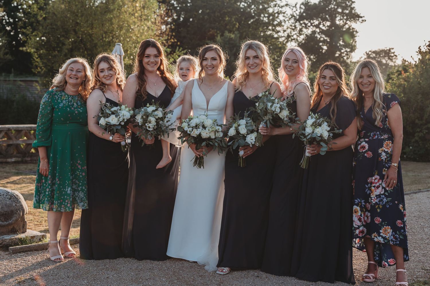 A bridal party poses together outdoors at sunset wearing mix of dark gowns and floral dresses while holding bouquets.