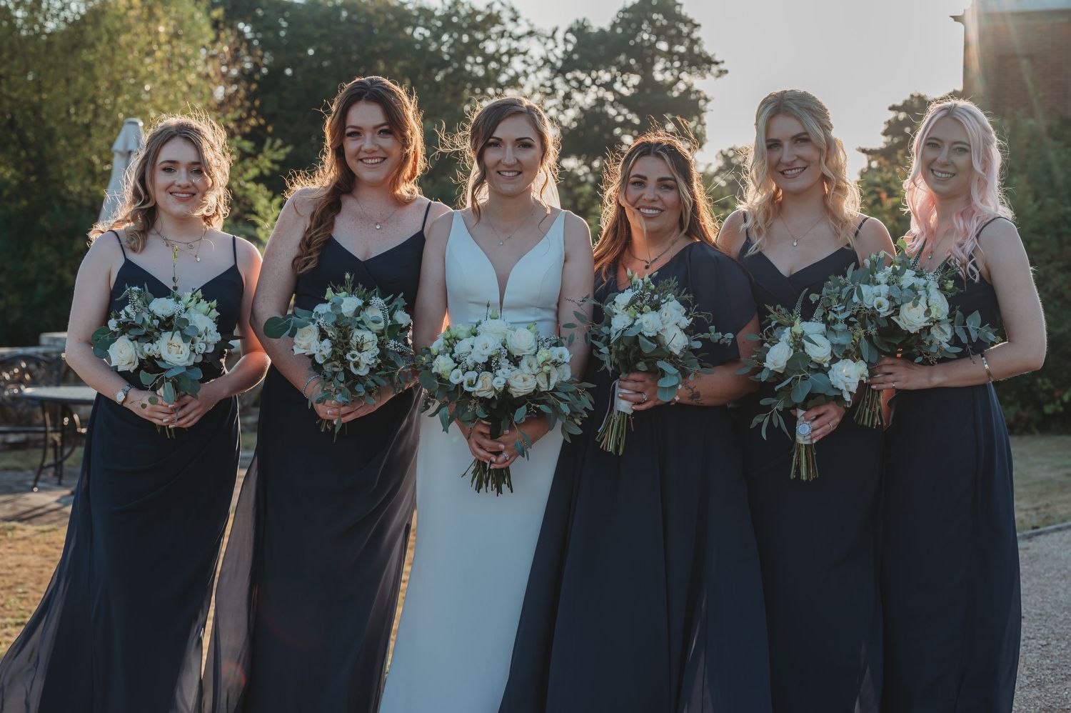A wedding party in navy blue bridesmaid dresses and a light blue bridal gown hold bouquets outdoors at sunset.