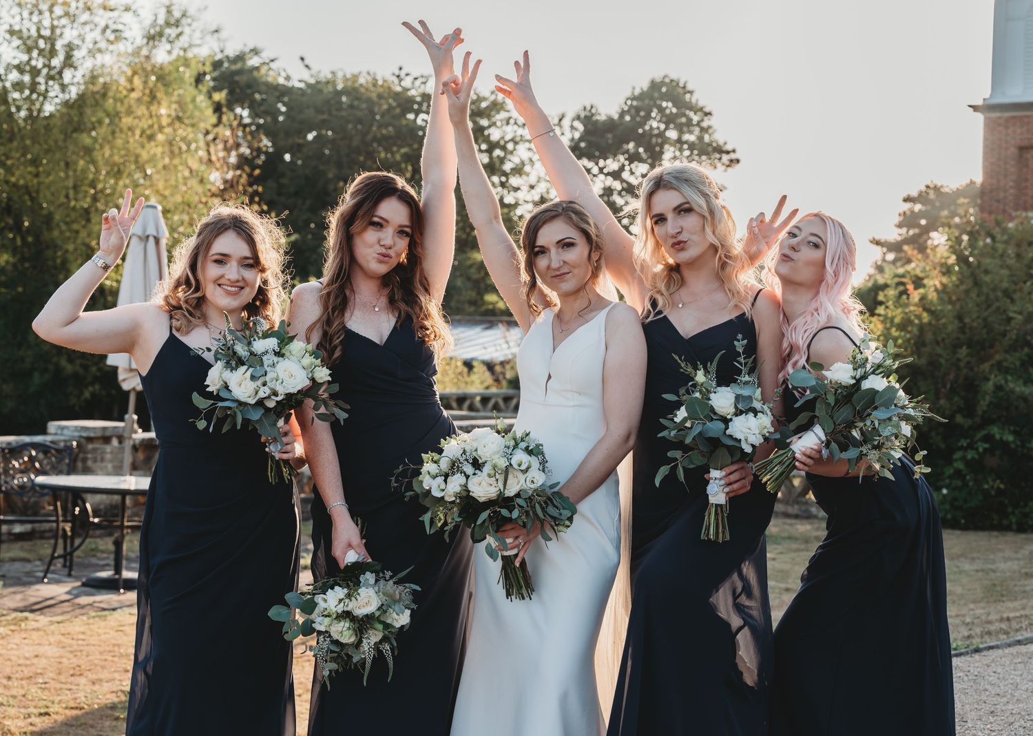 Five figures in formal attire hold bouquets for wedding celebration outdoors at sunset with trees in background.