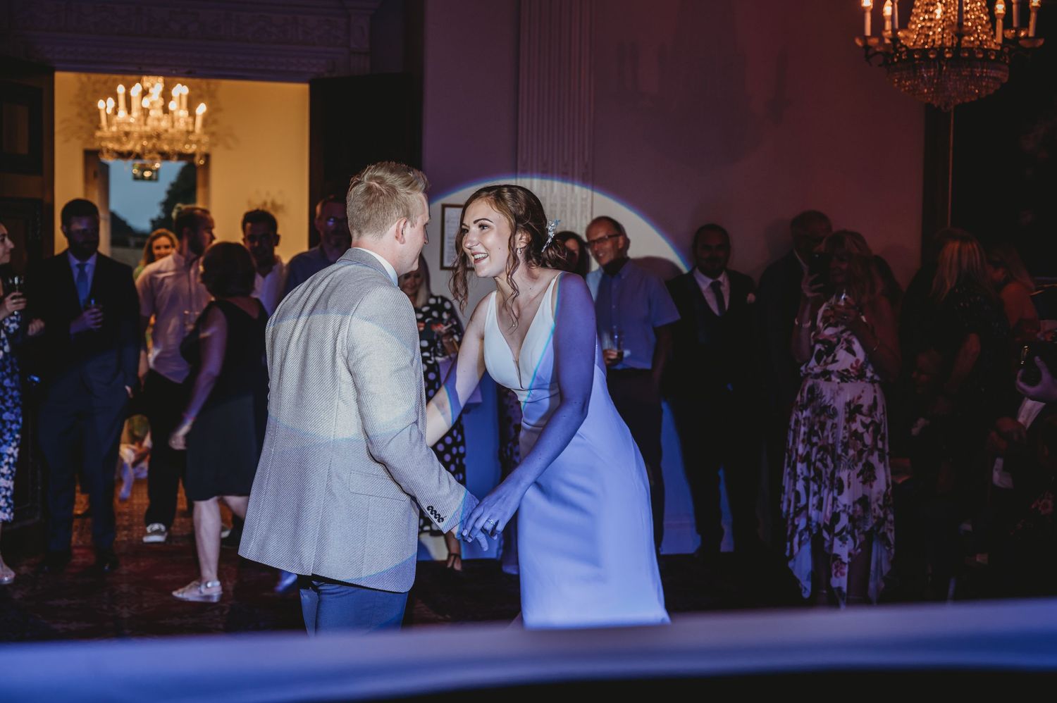 Couple shares an intimate dance moment on a dimly lit ballroom floor with guests looking on in the background.