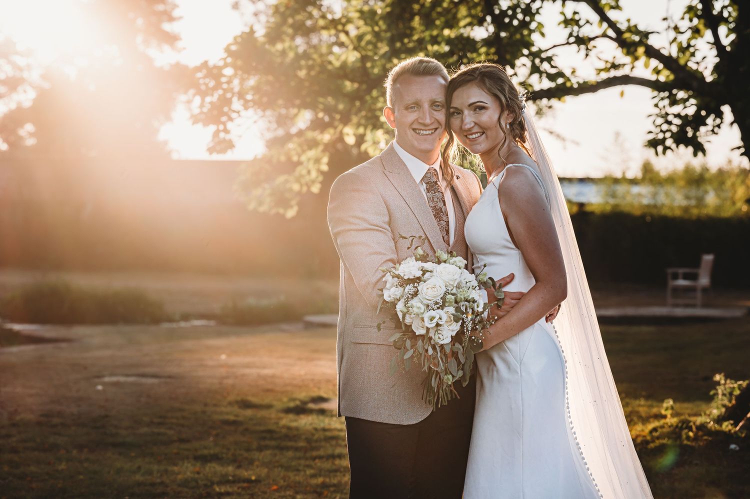 A couple in wedding attire pose outdoors at sunset holding a white floral bouquet against a sunlit countryside backdrop.