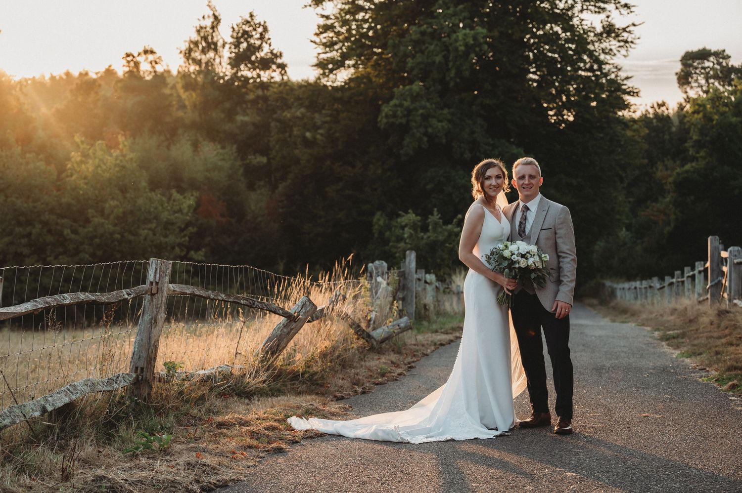 A couple shares an intimate sunset moment on a rural path lined with wooden fences and golden grass.
