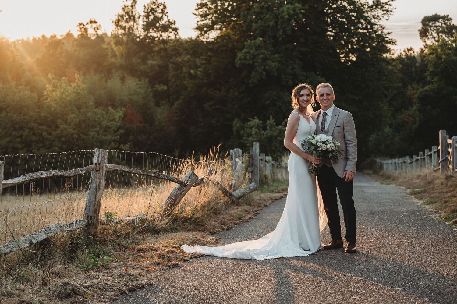 Couple in formal attire standing together on a country road at sunset with rustic wooden fence in background.