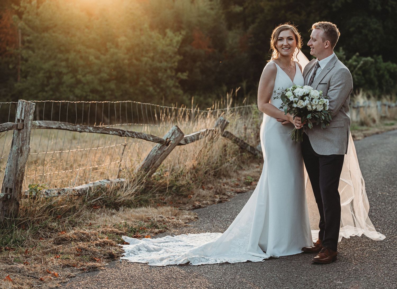 A couple in wedding attire stands together on a country road at sunset with rustic wooden fence in background.