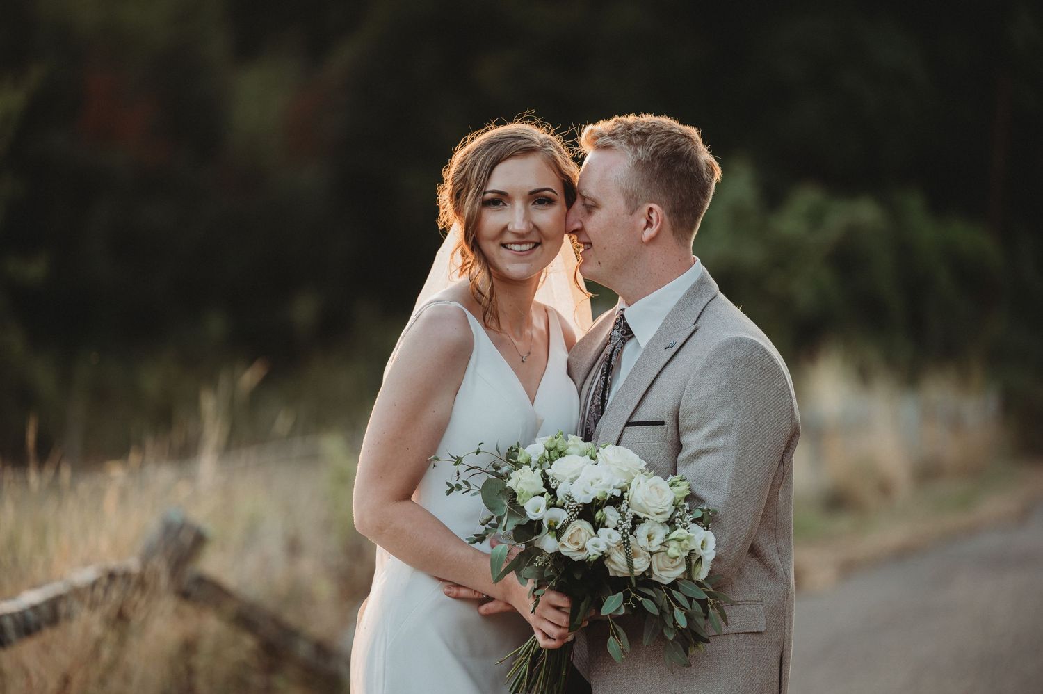 A couple embraces on a country road during golden hour while holding a white floral bouquet.