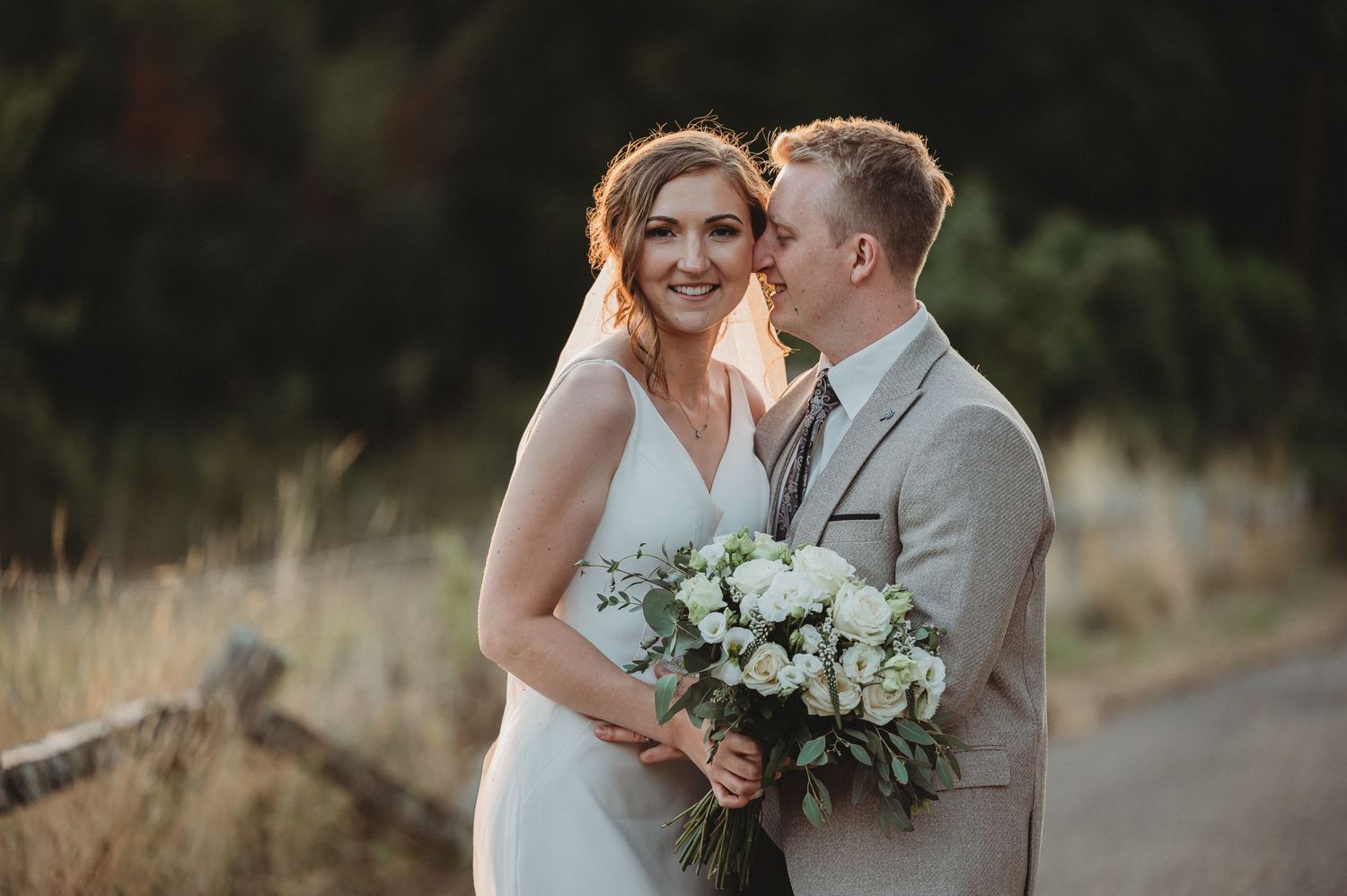A newlywed couple in wedding attire embrace on a path while holding a white and green floral bouquet.