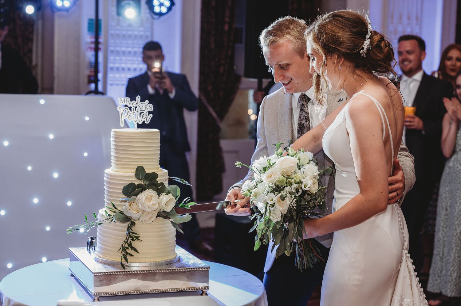 A couple cuts their elegant three-tiered white wedding cake during an indoor reception with twinkle lights.