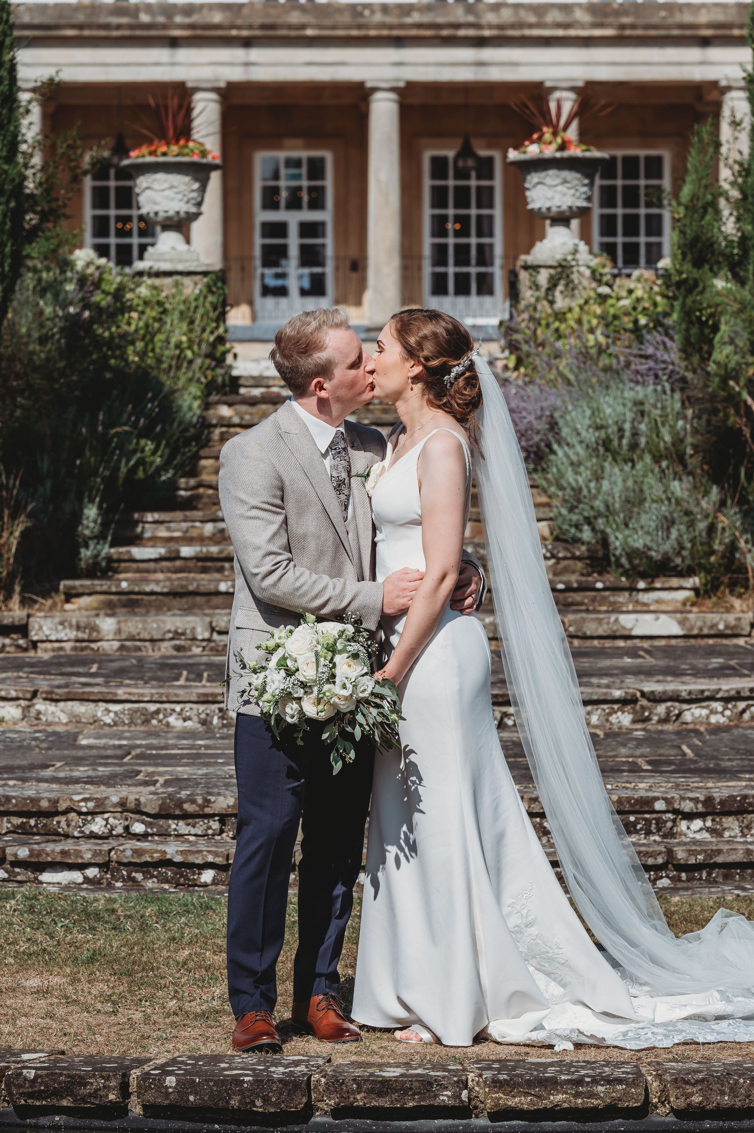 A newlywed couple sharing a romantic kiss on stone steps in front of a grand historic mansion with elegant columns.
