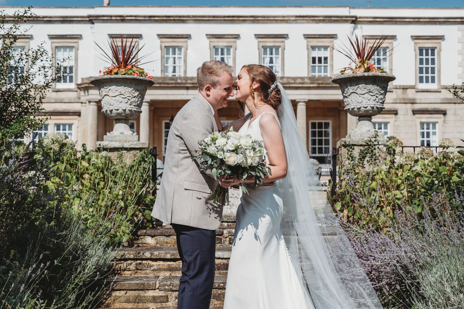 A rustic wedding couple shares an intimate moment in front of a grand manor house with ornate architecture and lavender gardens.