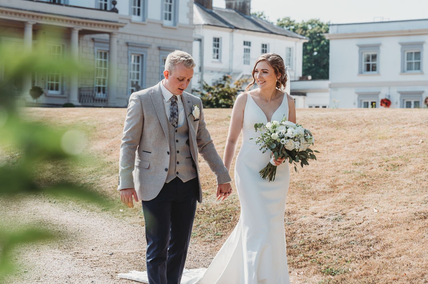 A wedding couple walks together on the grounds of an elegant country estate on a sunny summer day.