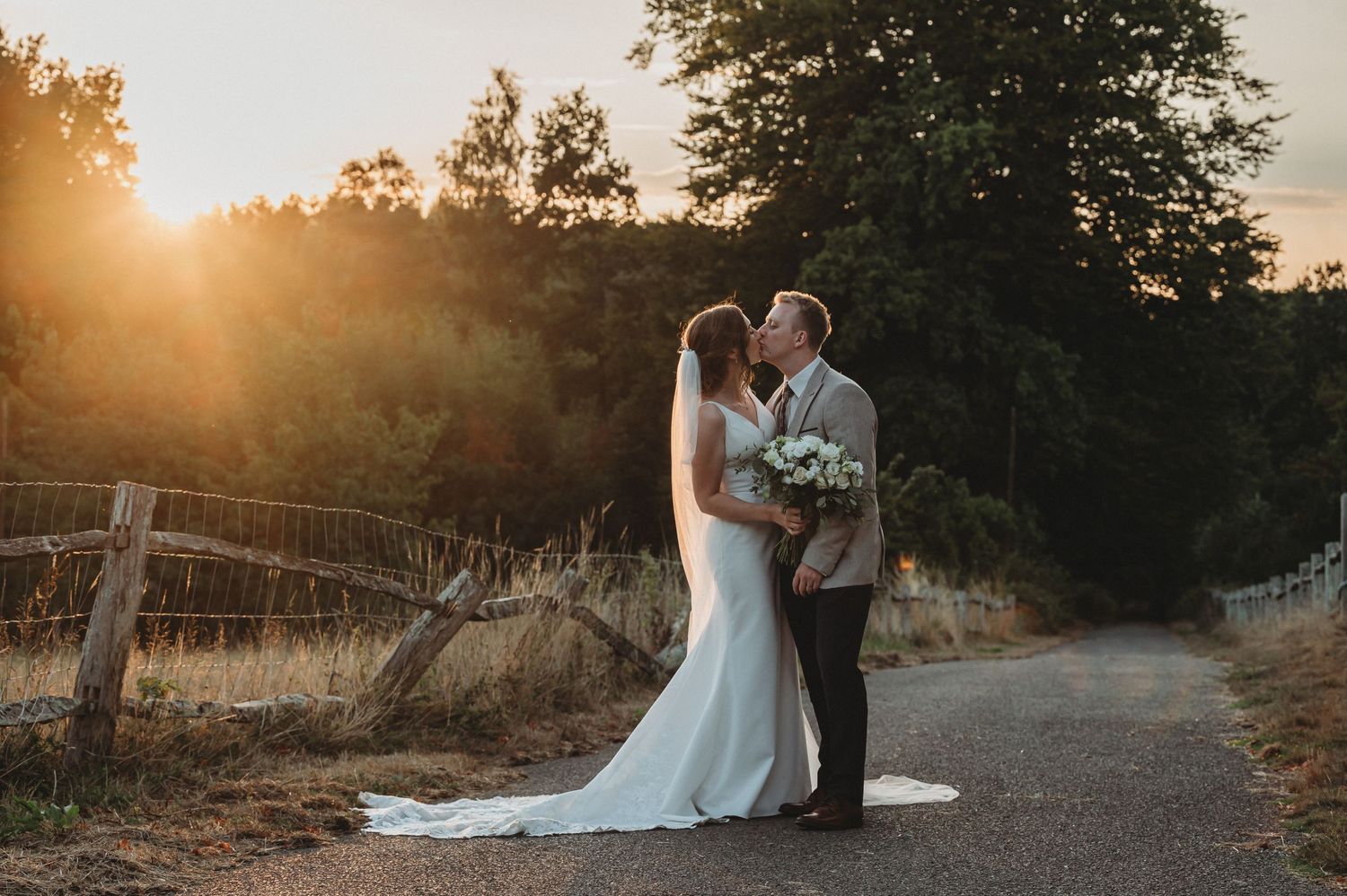 A romantic sunset wedding photo with a couple embracing on a rustic dirt path beside a wooden fence.