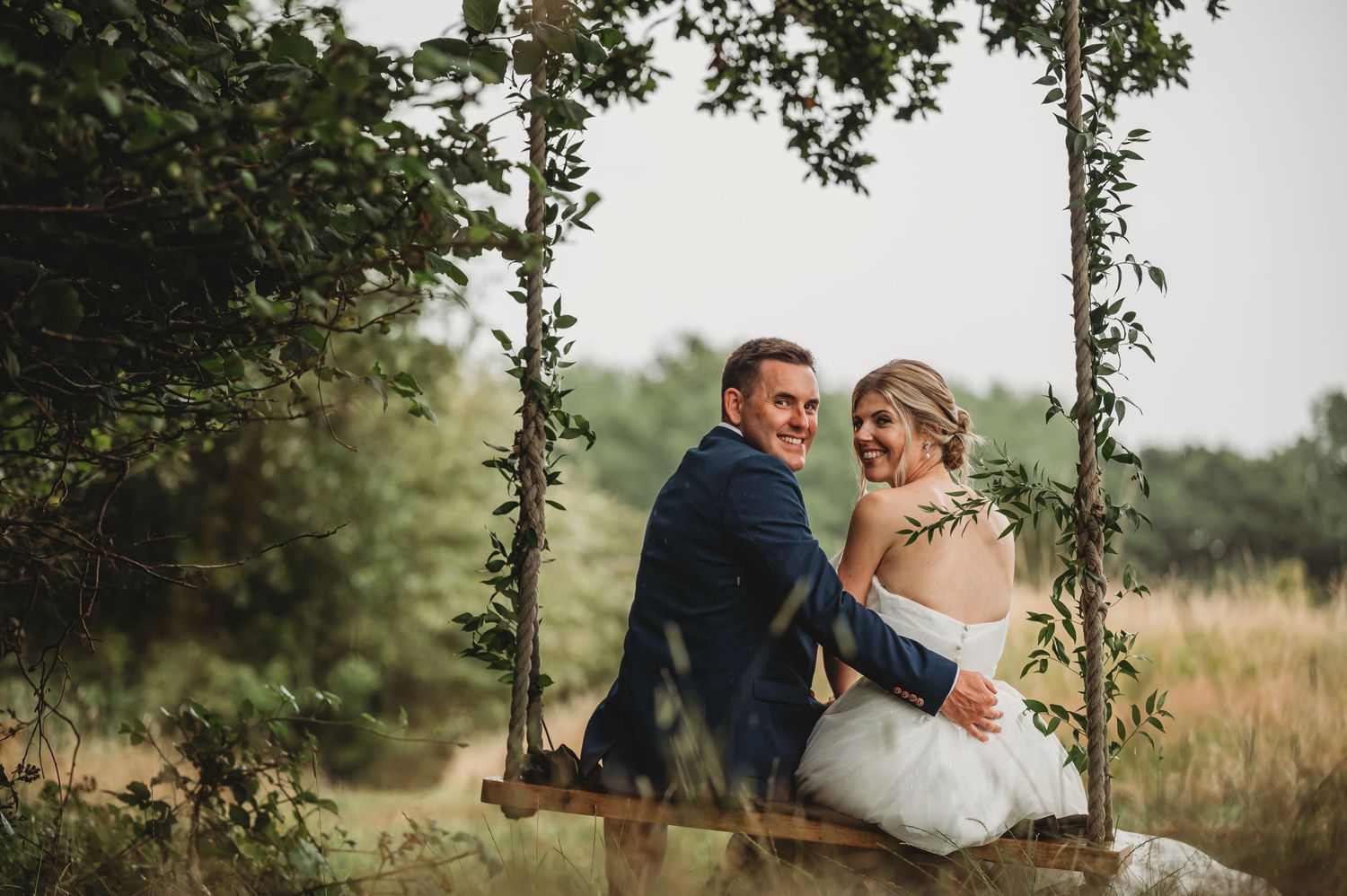 A couple sits together on a rustic wooden swing adorned with greenery in a dreamy outdoor setting.