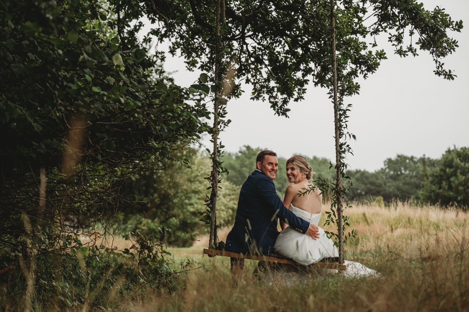 A couple sits together on a wooden swing surrounded by tall grass and trees in a natural outdoor setting.