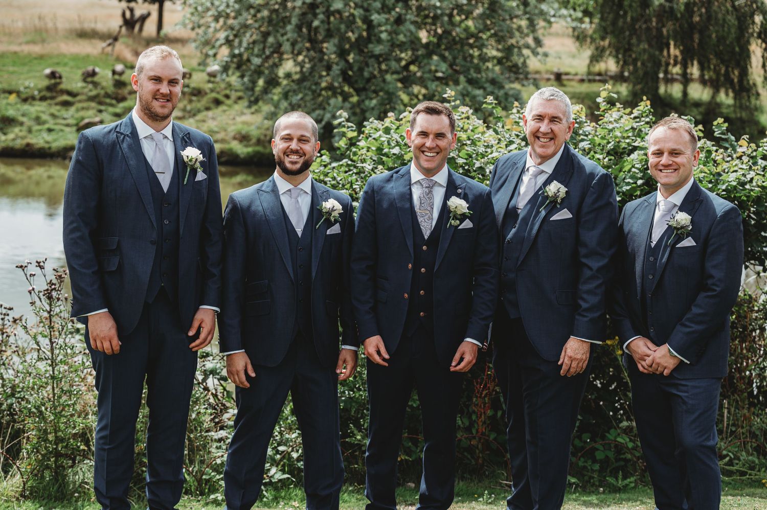 Groomsmen in navy suits pose together by garden pond.