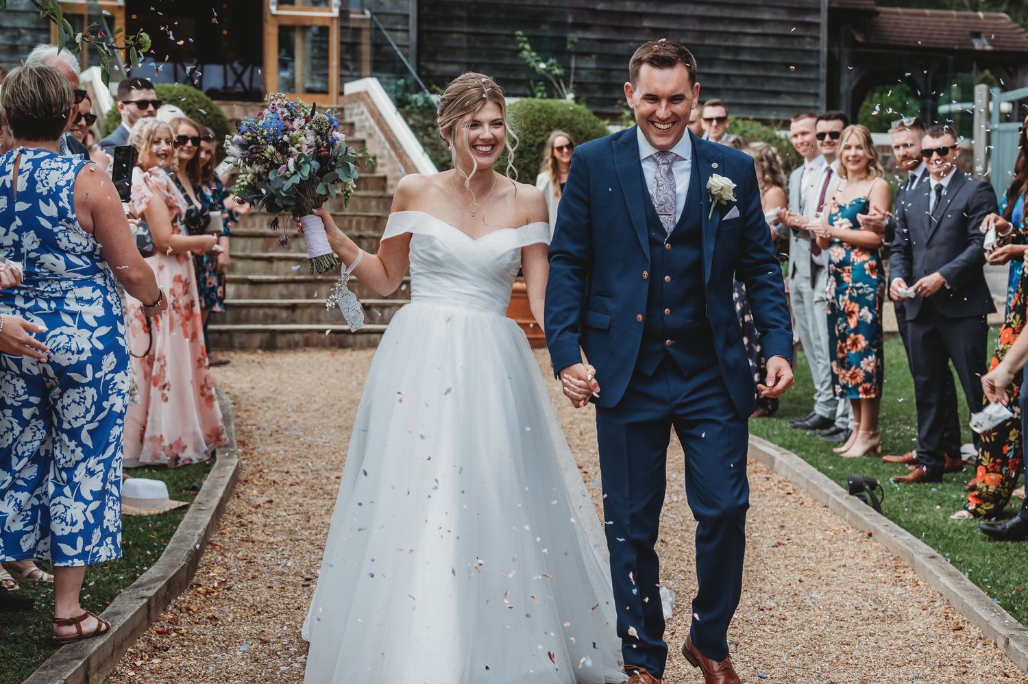 Newlyweds walk down a garden path lined with wedding guests while the bride wears a sparkling off-shoulder gown.