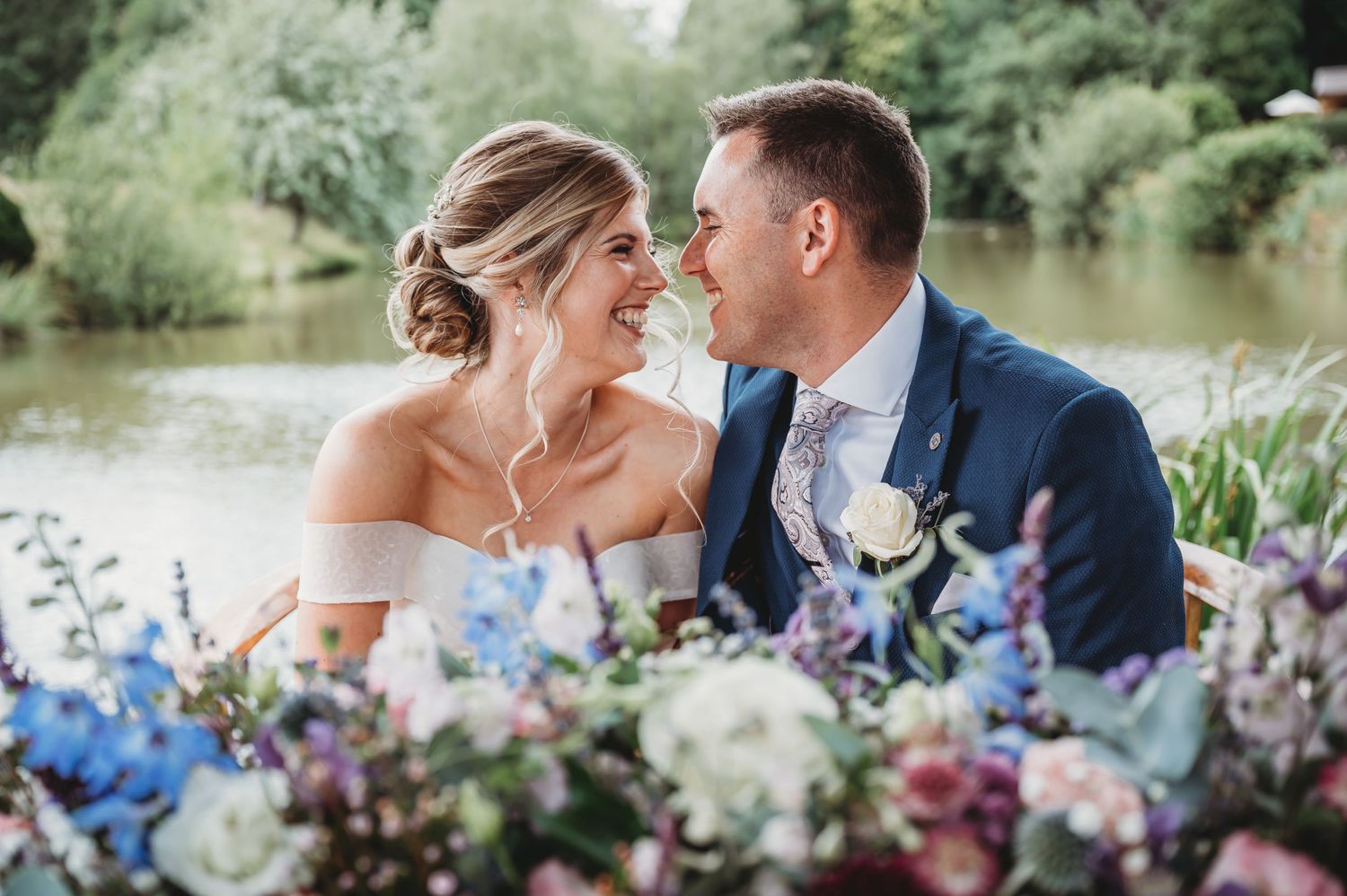 A romantic wedding couple embraces by a lakeside with elegant blue and white floral decorations in the foreground.