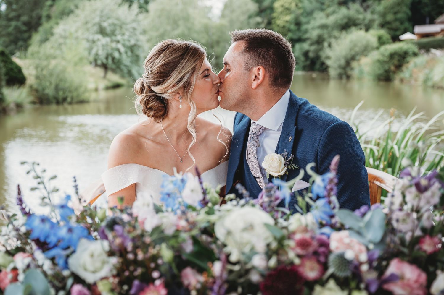 Romantic wedding couple shares a kiss by a lake with colorful blue and white floral arrangements in the foreground.