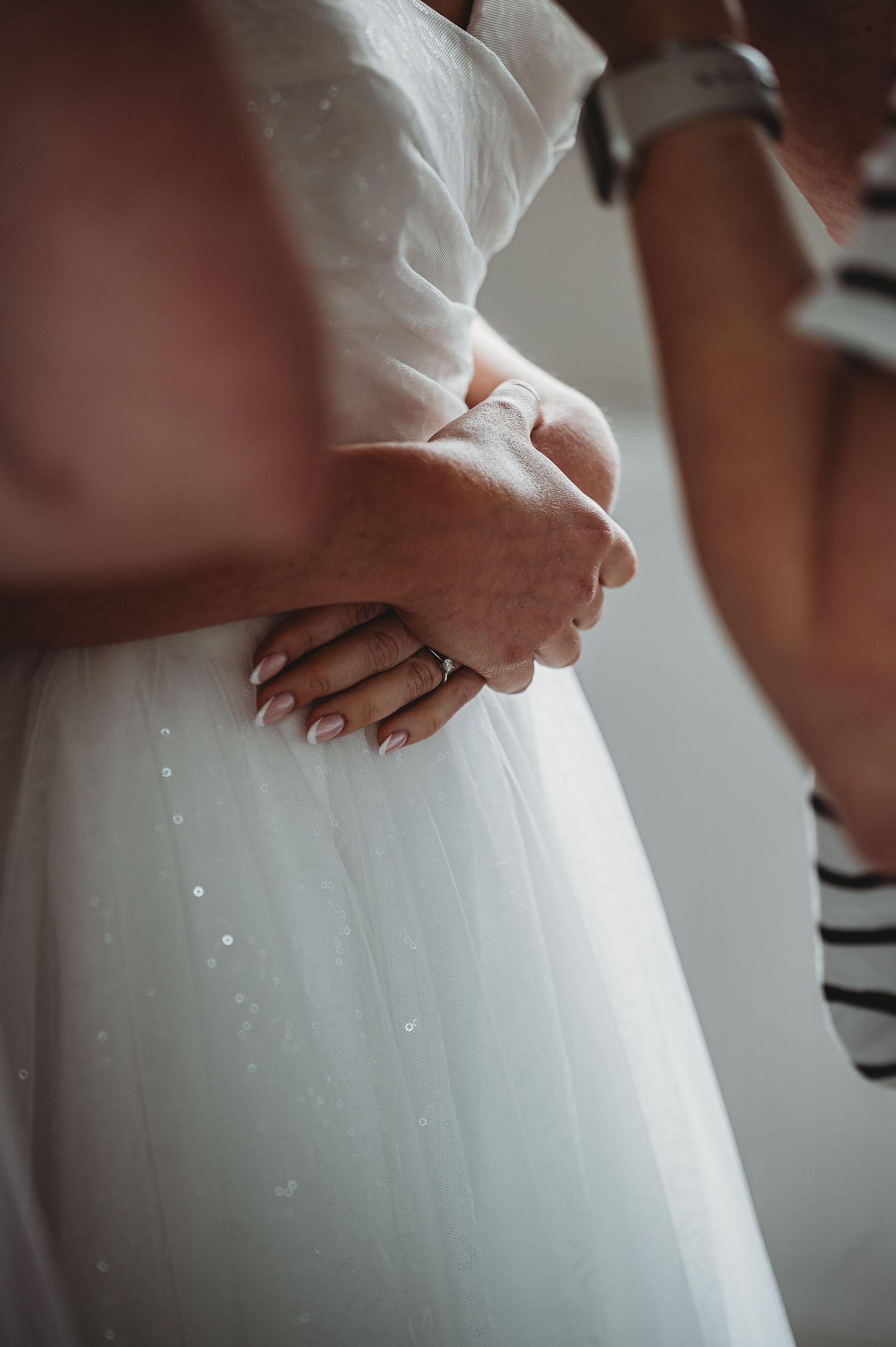 Close-up view of hands clasped over the waist of a white wedding dress with delicate sparkles and tulle fabric.