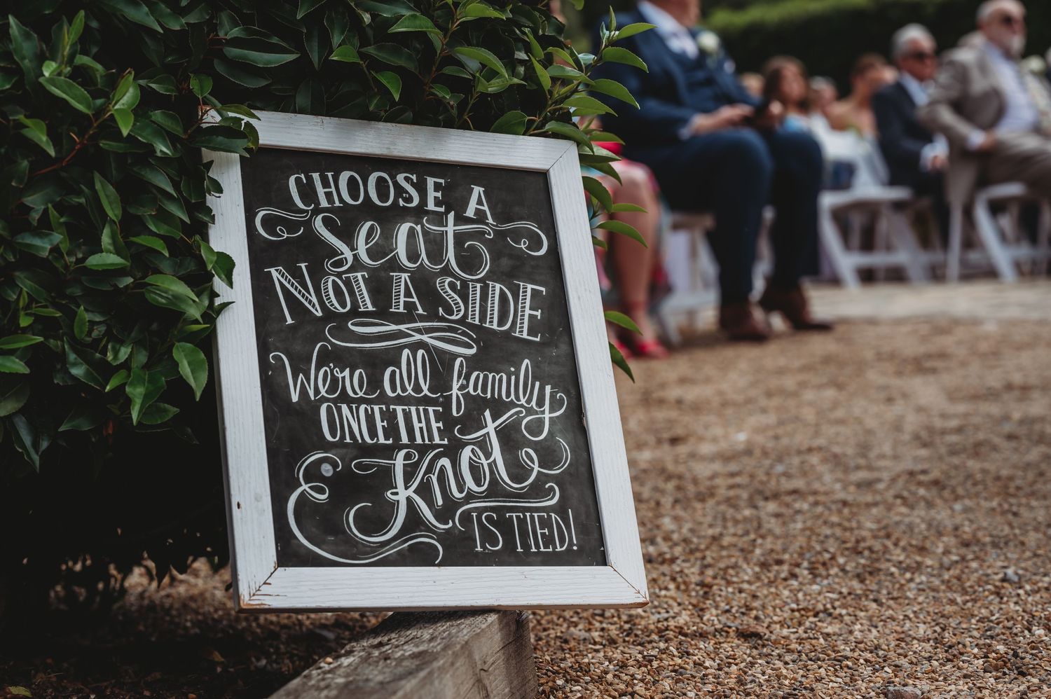A rustic chalkboard wedding sign that reads 'Choose a seat not a side' placed near greenery at an outdoor ceremony.