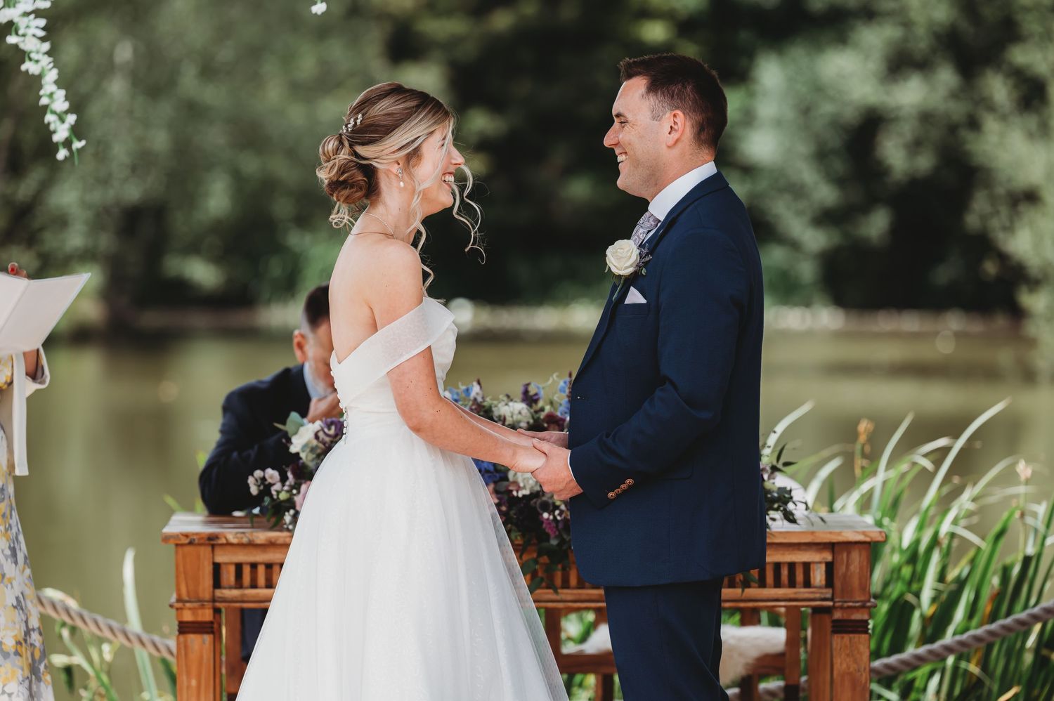 Wedding couple exchanging vows by a pond under a wooden arch decorated with flowers.