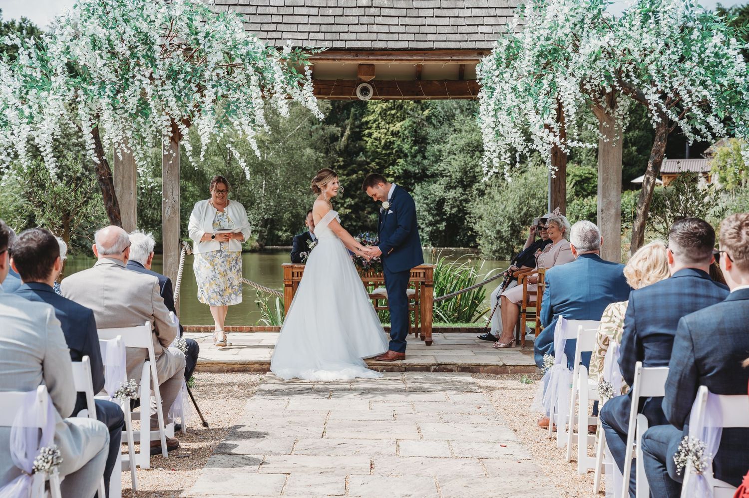 Outdoor wedding ceremony with guests seated under a wooden pergola draped with cascading white flowers.