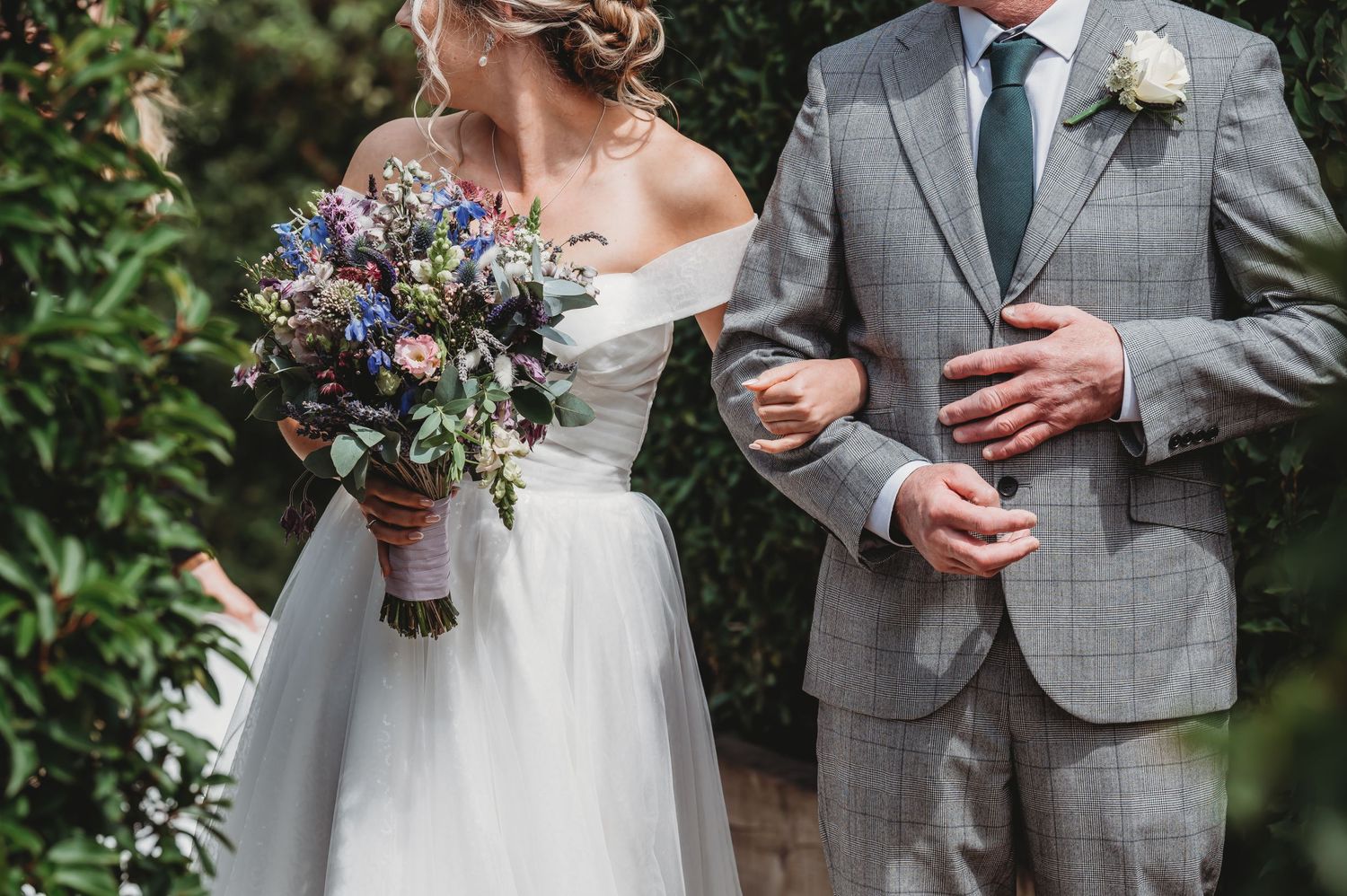 A couple in wedding attire holds an elegant wildflower bouquet featuring purple and blue blooms against a garden backdrop.