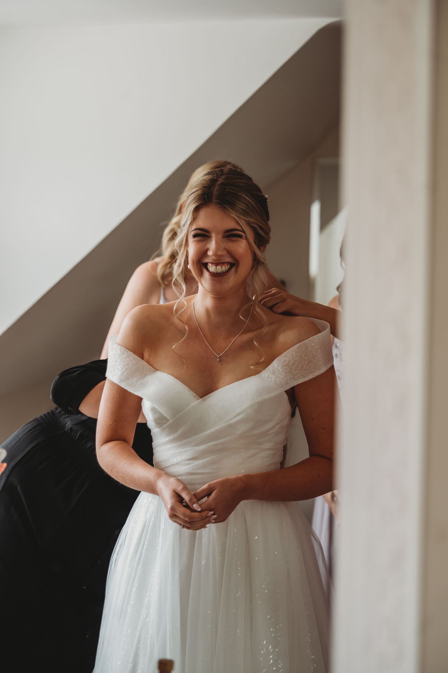 A bride wearing an elegant off-shoulder white wedding gown smiles joyfully while getting ready.