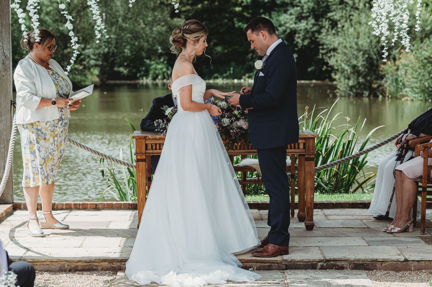 A lakeside wedding ceremony on a wooden platform with a bride in white gown and groom in navy suit exchanging vows.