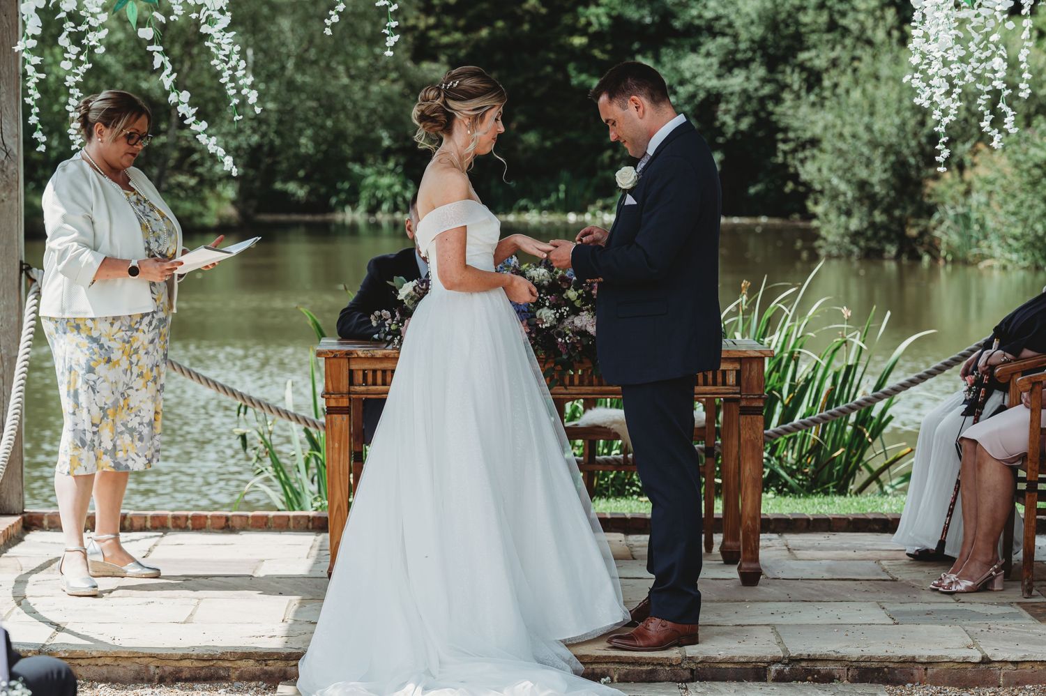 An outdoor wedding ceremony taking place on a dock by a pond with hanging string lights and greenery in the background.