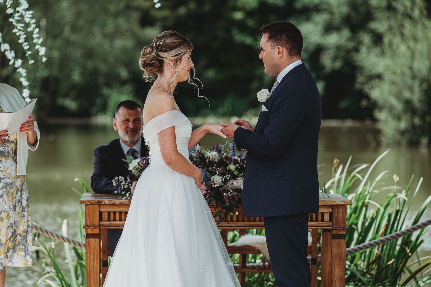 A wedding ceremony takes place on a deck by a lake as the couple exchanges rings surrounded by greenery.