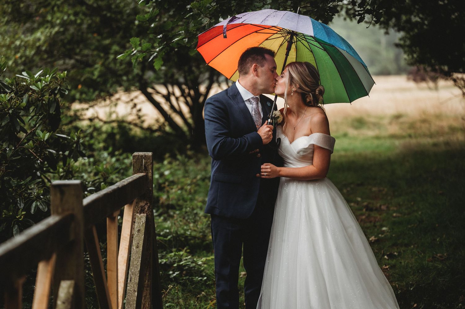 Romantic wedding couple shares intimate moment under colorful rainbow umbrella in outdoor garden setting.