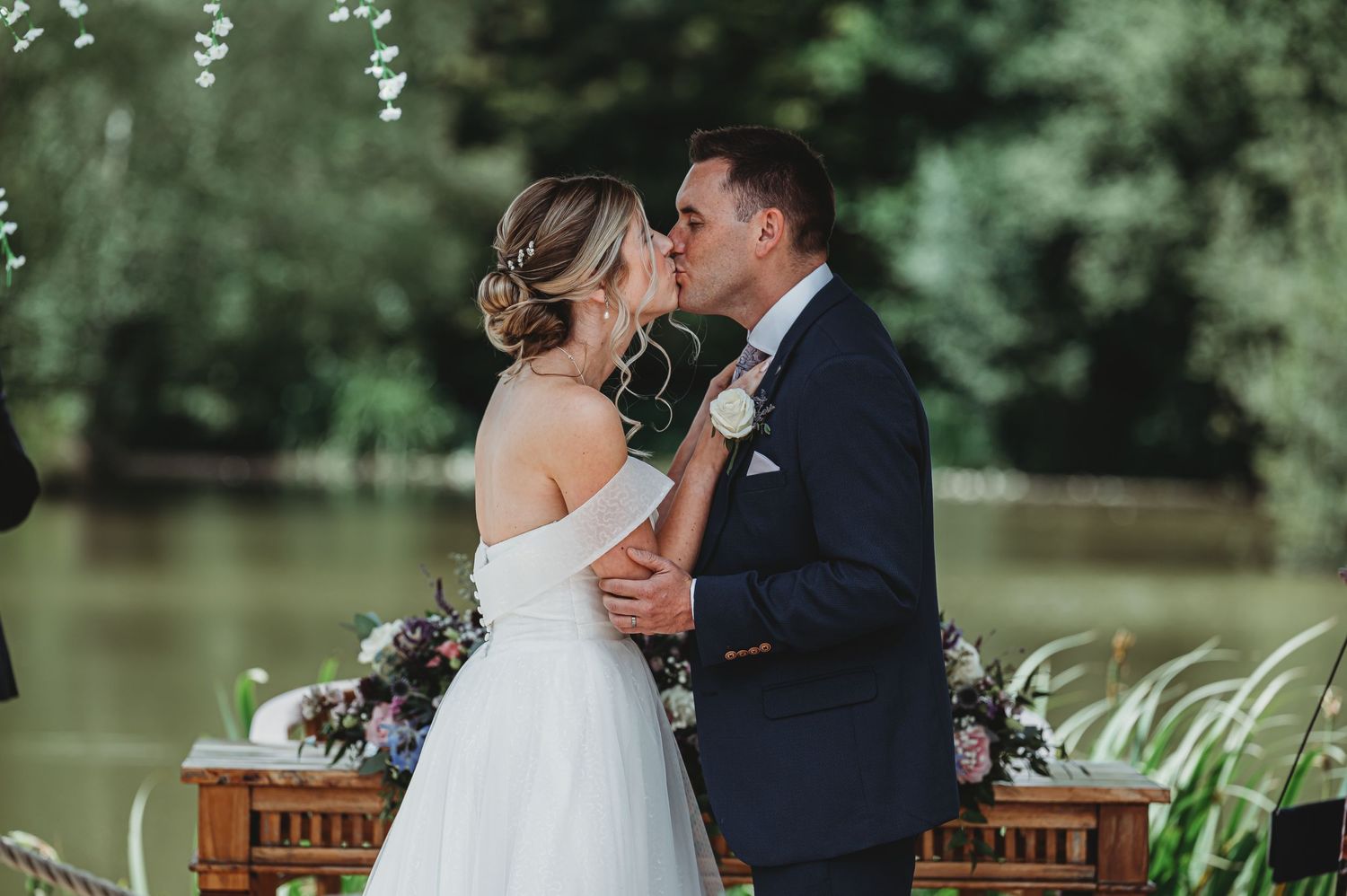 A romantic outdoor wedding ceremony with a couple kissing by a lake with wooden furniture and floral decorations.