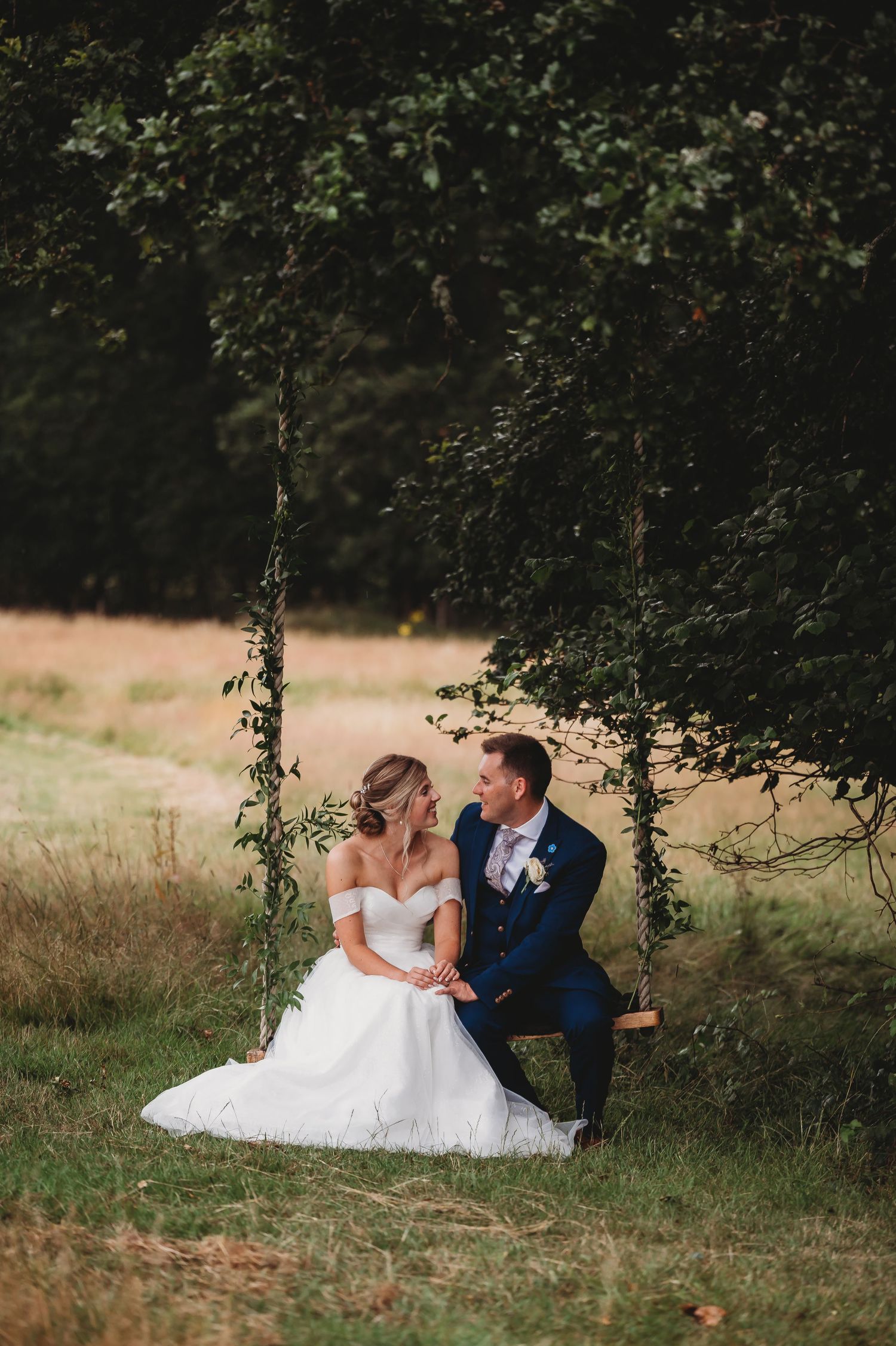 A couple sits together on a rustic wooden swing beneath overhanging tree branches in a romantic outdoor setting.