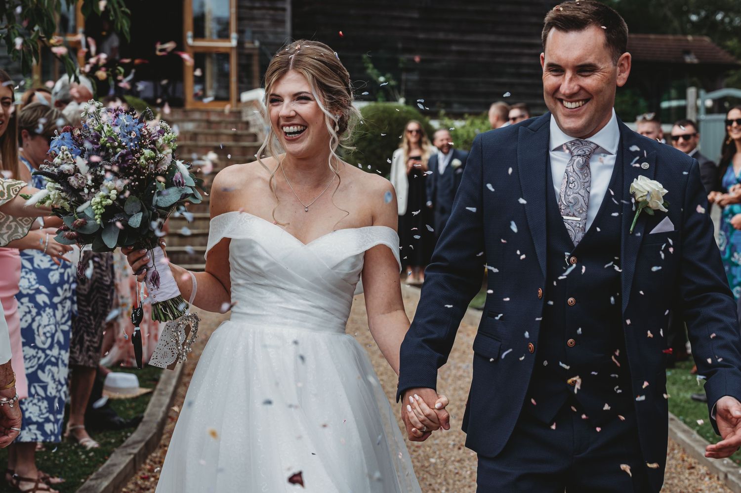 A joyful newlywed couple walks hand in hand while guests shower them with confetti outside a brick building.