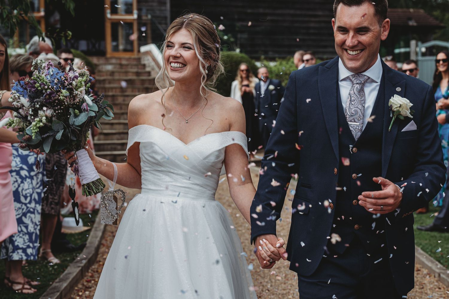 Two newlyweds walk hand in hand while confetti falls around them at an outdoor wedding ceremony.