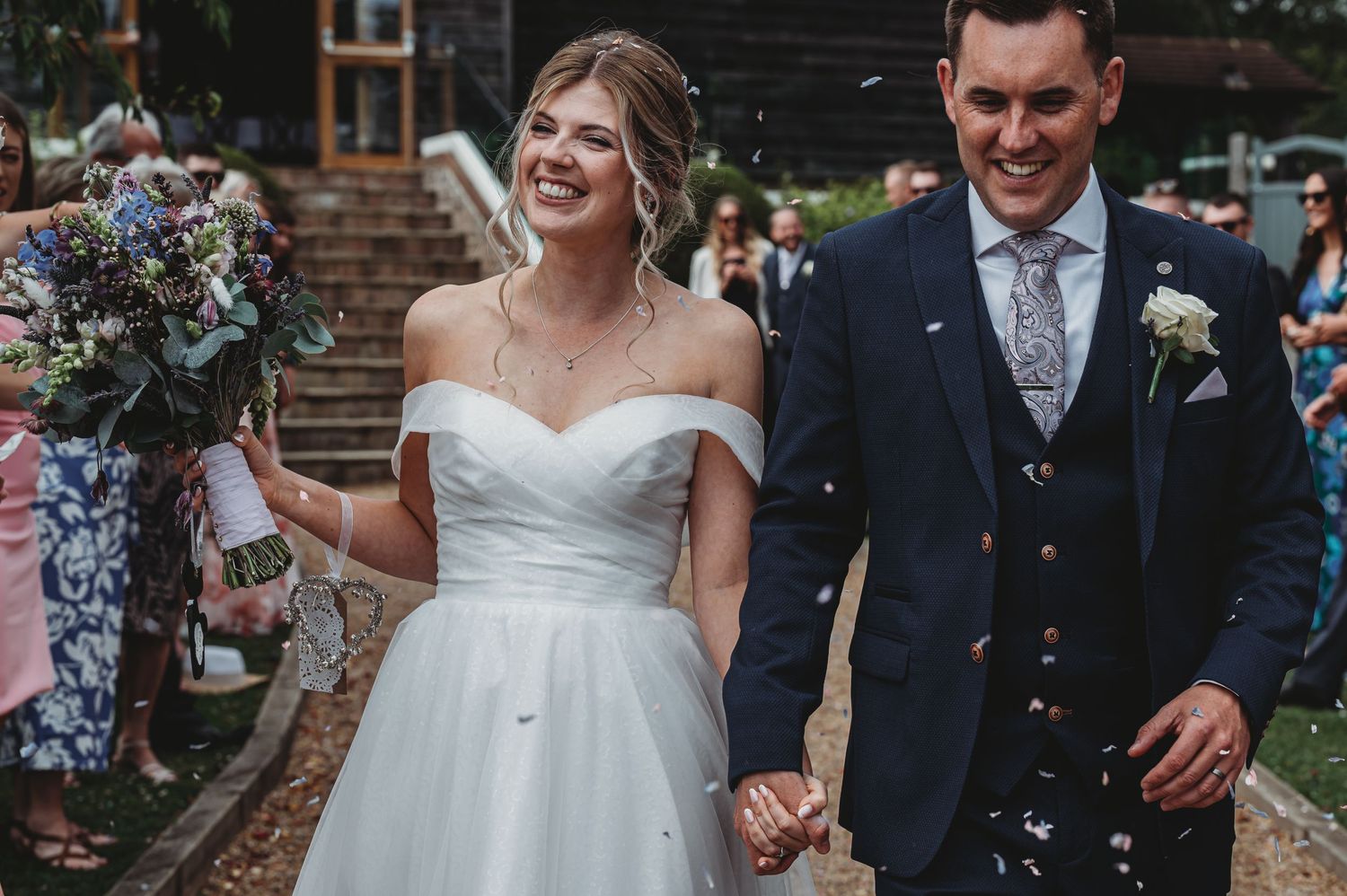 Joyful newlyweds walk through shower of confetti after wedding ceremony with bride in white dress and groom in navy suit.