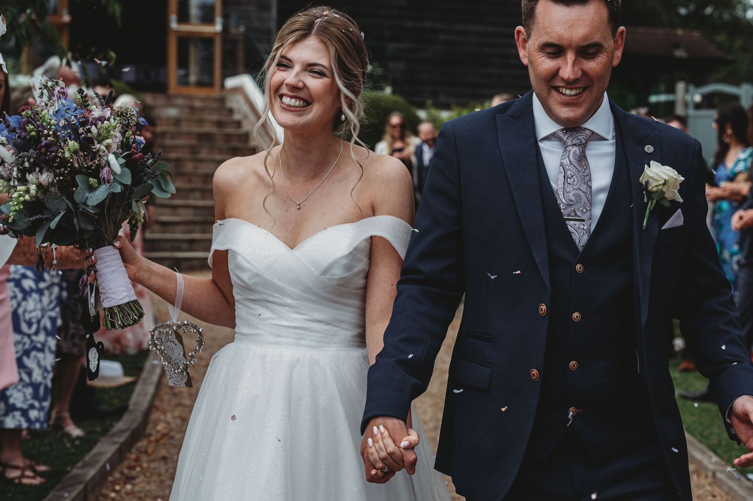A couple walking hand in hand after their wedding ceremony, bride in white off-shoulder dress and groom in navy suit.