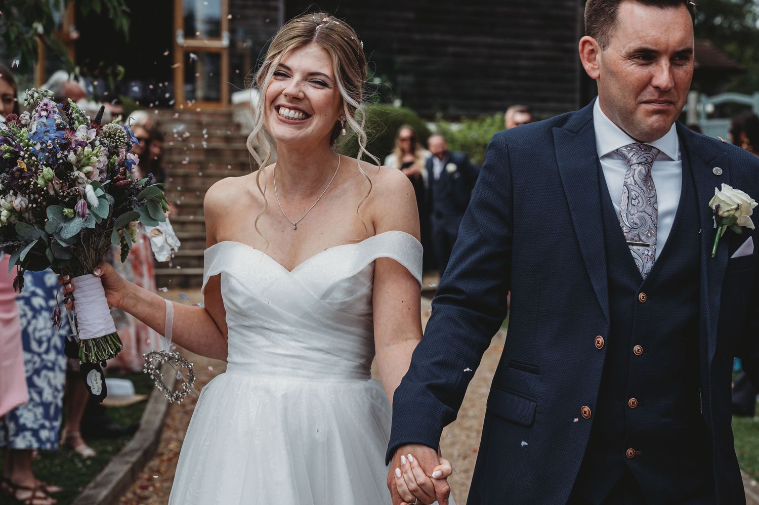 A bride in an off-shoulder white dress holds a bouquet while walking with groom in navy suit at outdoor wedding.