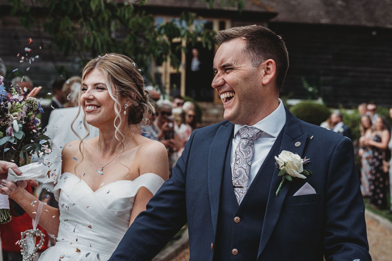 Happy newlyweds share a joyful moment with guests throwing confetti during outdoor wedding celebration.