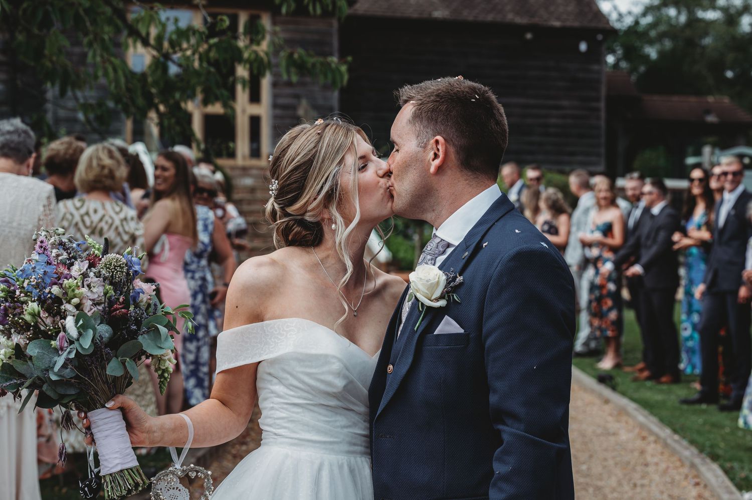 Newlyweds share a kiss at outdoor wedding reception while guests look on from the garden background.