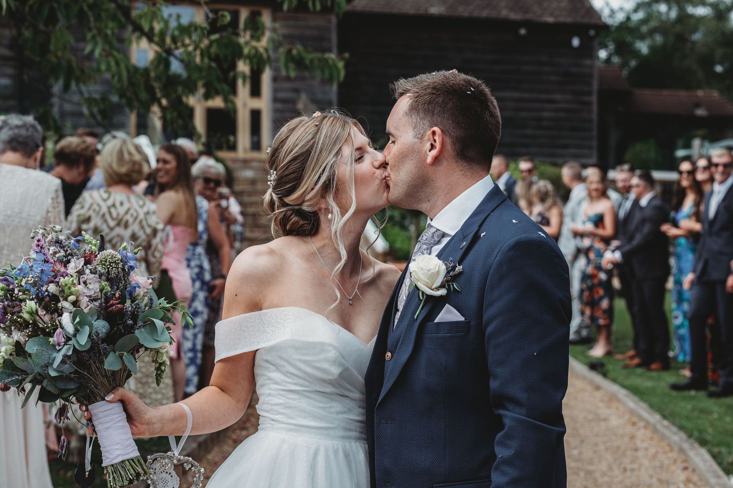 Happy newlyweds share a kiss at their outdoor wedding ceremony as guests look on in the background.
