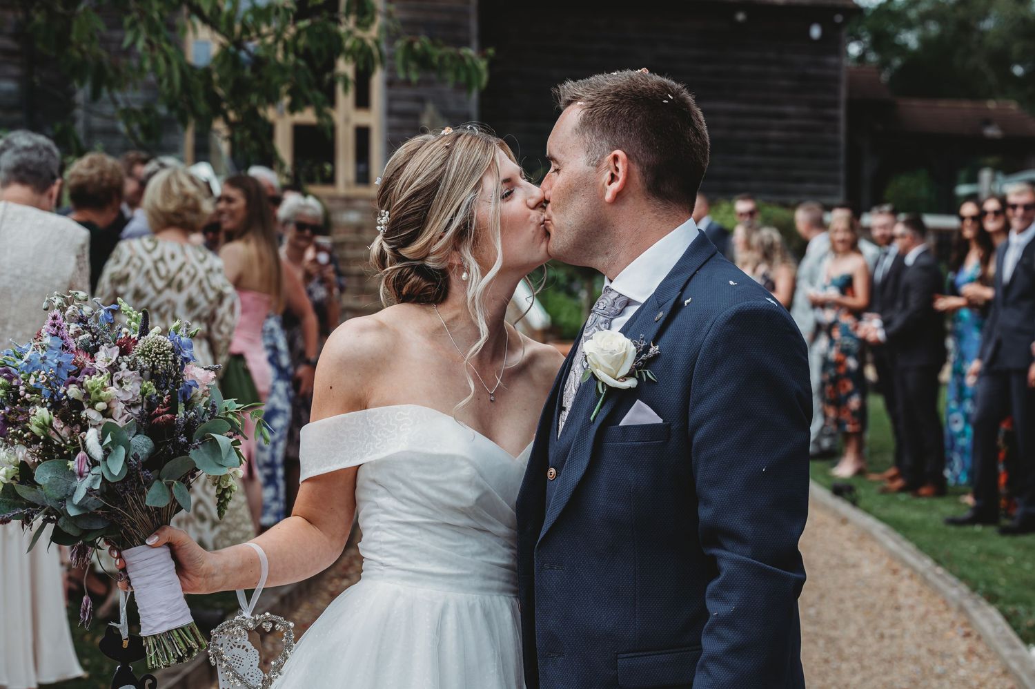 A romantic outdoor wedding moment showing a couple sharing a kiss as guests look on from the garden path.