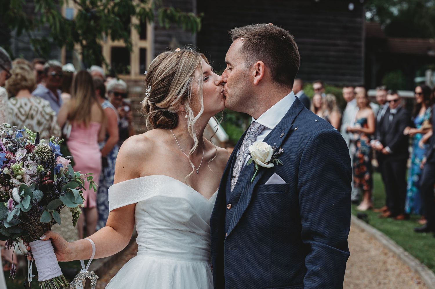 A romantic kiss at an outdoor garden wedding reception as guests look on among floral decorations.