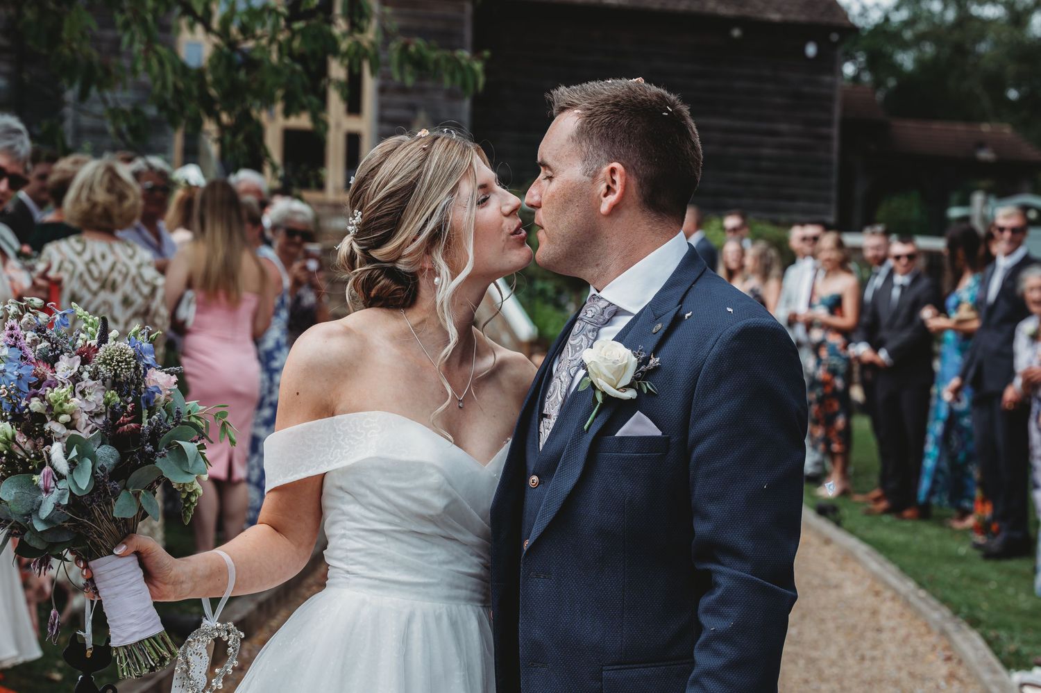 Couple shares a romantic kiss at outdoor garden wedding ceremony while holding floral bouquet.